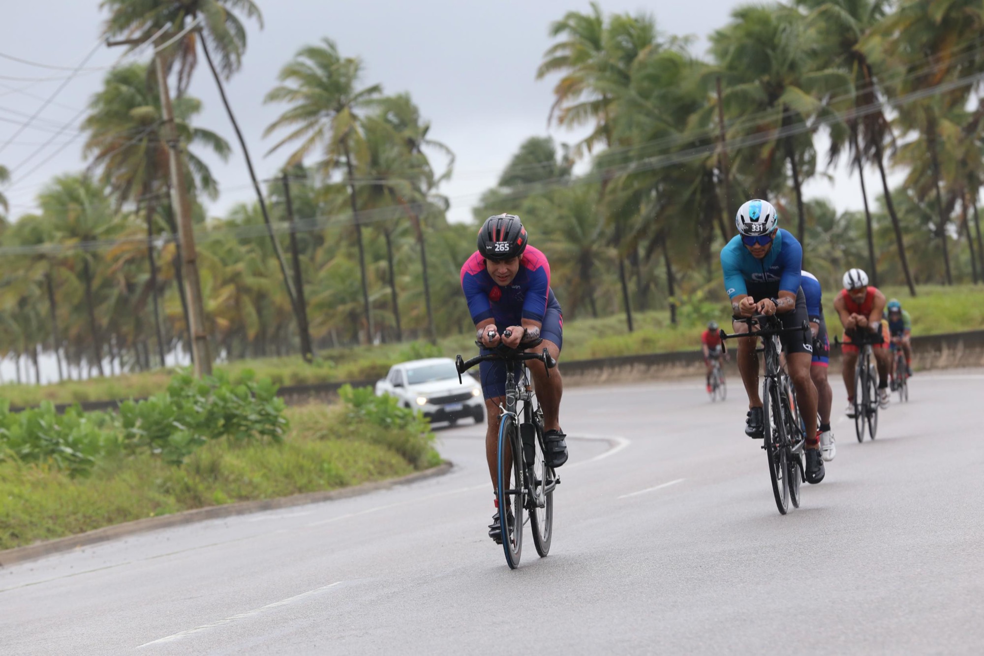 Carlos Endrigo na etapa de ciclismo do IRONMAN 70.3 Maceió, liderando um pelotão numa estrada à beira-mar com coqueiros